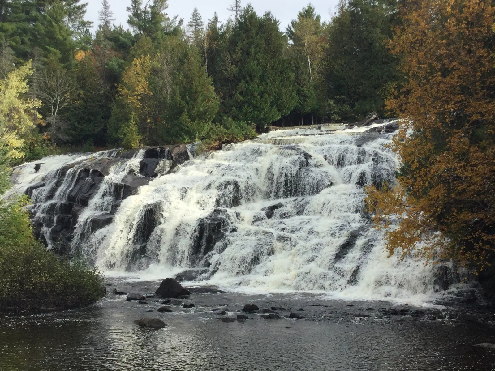 The wide tiered curtain of Bond Falls in autumn, whitewater stepping down dark rock the full width of the river.