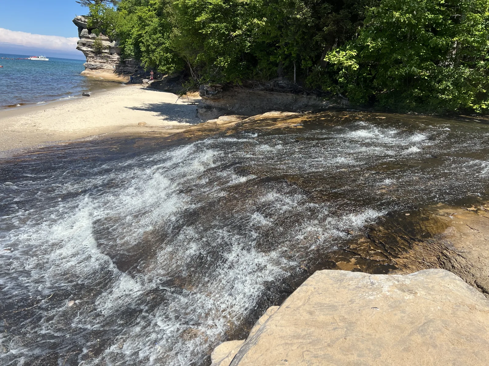 Chapel Beach Falls sliding across a wide sandstone shelf into Lake Superior at Chapel Beach, with Chapel Rock and the beach behind it.