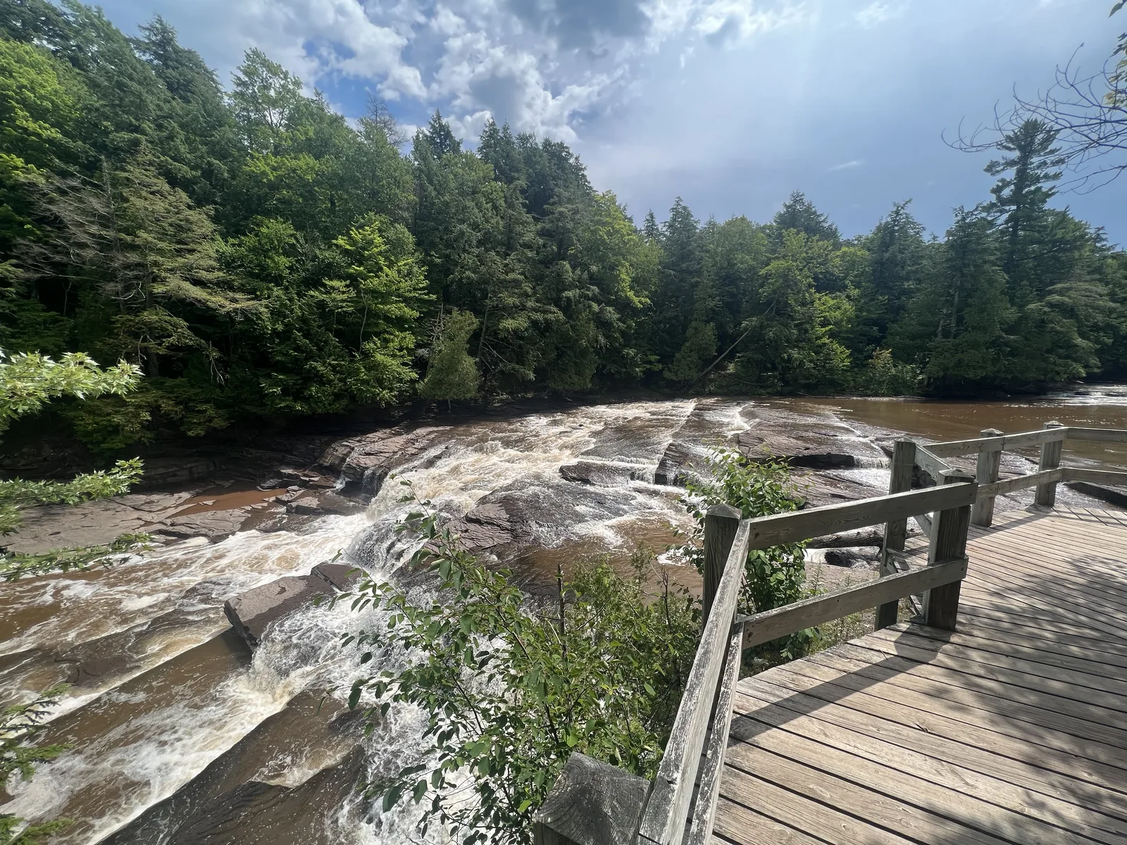 Manido Falls wide cascade pouring over reddish rock shelves on the Presque Isle River, viewed from a wooden boardwalk.
